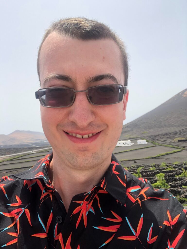 Matthew is taking a selfie, from his shoulders upwards and is smiling at the camera. He is wearing black sunglasses and a black shirt with vibrant red and blue leaves on it. The background is lush, mountainous and the sun is shining.