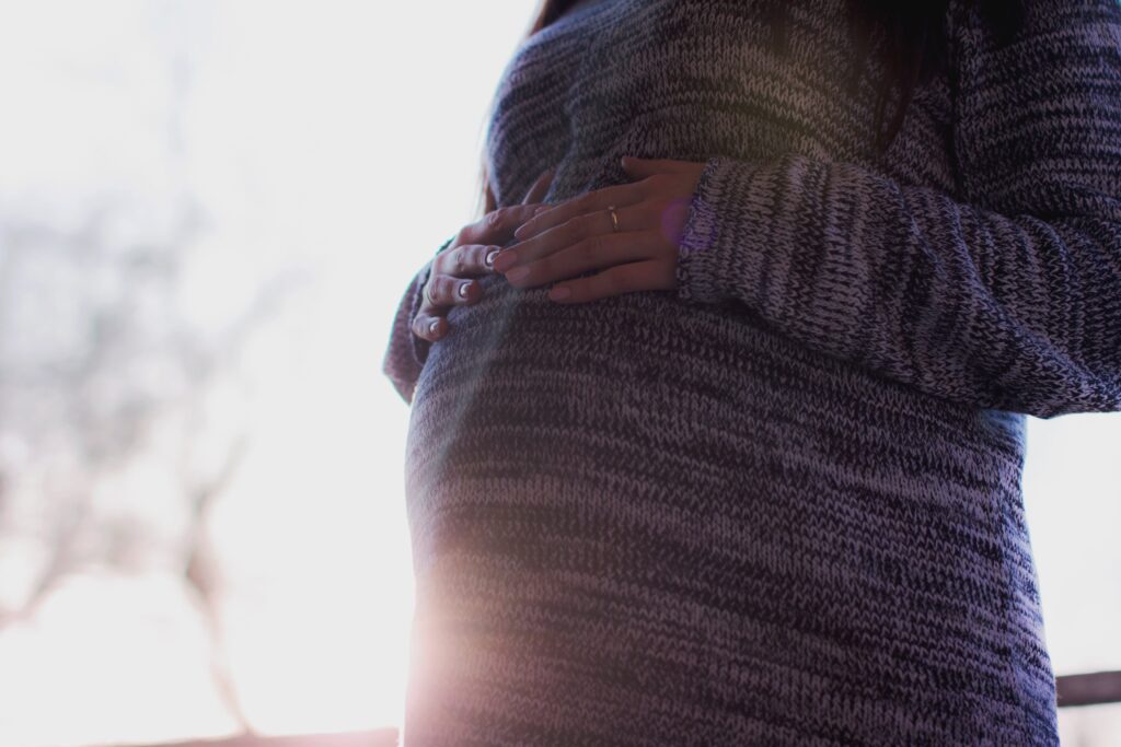 A pregnant woman wearing a marl grey jumper, resting her hands on her stomach