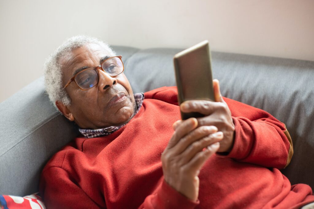 An elderly man laying on a sofa, wearing a red jumper. He is wearing glasses and is looking up at his mobile phone.