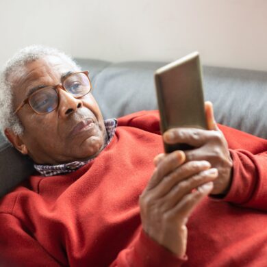An elderly man laying on a sofa, wearing a red jumper. He is wearing glasses and is looking up at his mobile phone.