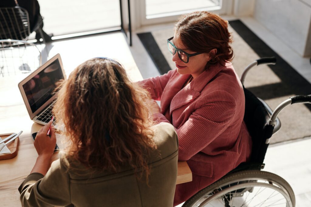 A cropped photo of two women sat chatting in front of a laptop. One woman is in a wheelchair and wears a red jacket and the other wears a khaki blouse.