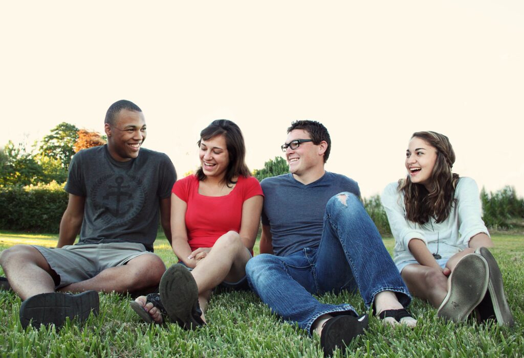 A group consisting of two women and two men sat on green grass. Their legs are in front of them and they are laughing.
