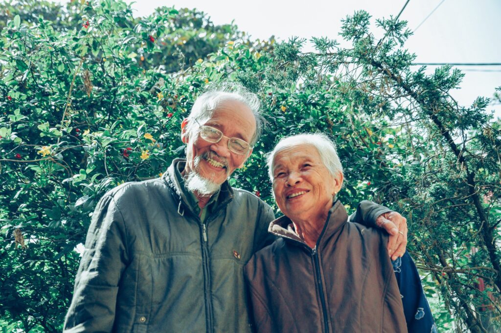 Smiling man and woman wearing jackets. The man has his arm around the woman and they are stood in front of green trees.