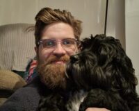 James is smiling at the camera with his dog to the right. The photo is cropped to show the top of his arms upwards. He is wearing a black jumper, has ginger hair and a beard. He is also wearing glasses. His dog is black and curly, with a white patch on its chest and a few white whiskers.