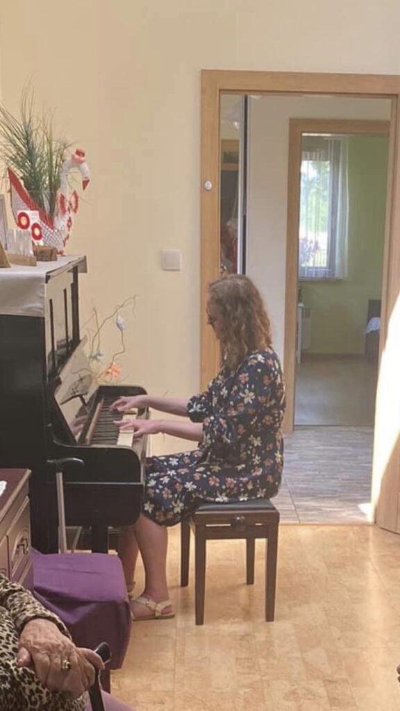 Dominika is sat on a brown chair, playing a piano. She is wearing a long sleeve, mid-length, navy, floral dress and white sandals. She has wavy hair and is looking down at the keys.