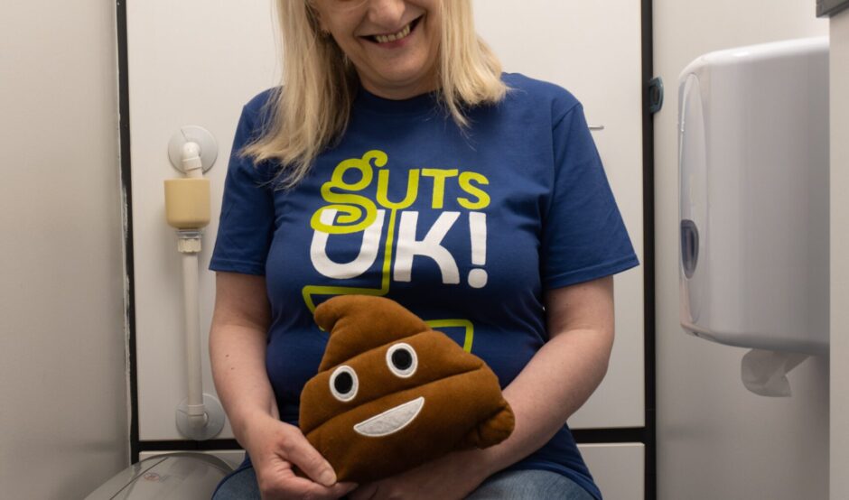 Julie is sat on the toilet wearing dark blue denim jeans (that are pulled up!) and a Guts UK t-shirt. She is holding a poo emoji cushion and is smiling at the camera.
