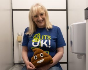 Julie is sat on the toilet wearing dark blue denim jeans (that are pulled up!) and a Guts UK t-shirt. She is holding a poo emoji cushion and is smiling at the camera.