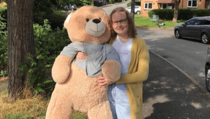 Dominika is wearing a white t-shirt, light denim jeans and a yellow cardigan. She also is wearing black rimmed glasses and is smiling at the camera, holding a light brown giant teddy bear.