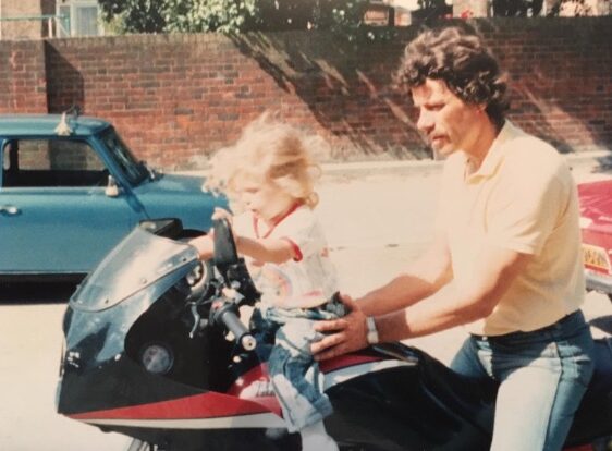 John in 1988, around the age of 36 to 37 is sat on his motorcycle seat while holding Charlotte as a child in front of him. 