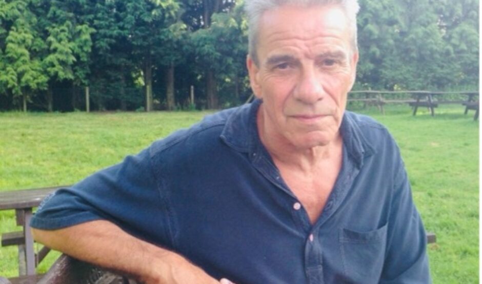John in 2013, aged 61, sat on a park bench wearing a blue navy shirt looking at the camera.
