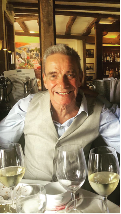 John in 2019 in a restaurant, sat with his glasses of white wine on the table and smiling at the camera.