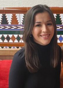 Sophie is looking at the camera smiling and has her hair down. She is wearing a black turtleneck long-sleeved top sat in front of a patterned tiled wall.