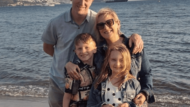 Sarah is standing on the beach with the sea waves behind her. She is standing next to her husband with his hand on her shoulder. She has her hands wrapped around her two children who are standing in front of her. All four of them are smiling at the camera.
