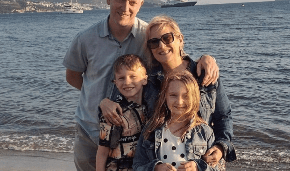 Sarah is standing on the beach with the sea waves behind her. She is standing next to her husband with his hand on her shoulder. She has her hands wrapped around her two children who are standing in front of her. All four of them are smiling at the camera.