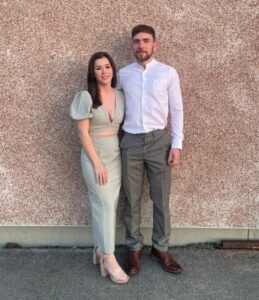 Sophie has her hair down and is wearing a pastel mint green full length dress with beige heels. Her partner has his right arm around her wearing a smart white long-sleeved shirt with formal gray trousers and brown leather Chelsea boots. Both are smiling at the camera in front of a brown wall.