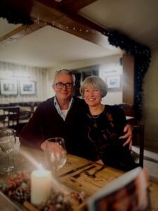 Geoff is sat smiling facing the camera in a rustic restaurant with his left arm wrapped around his wife. They are sat down behind a wooden table with two wine glasses and cutlery on top. Geoff is wearing his black long-sleeved polo top that is slightly unzipped in front.