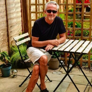 Geoff is sat outside on the patio surrounded by greenery smiling at the camera. He has his legs crossed and hands overlapping on one another while placed on the wooden table. He is wearing black sunglasses, black t shirt with beige shorts.
