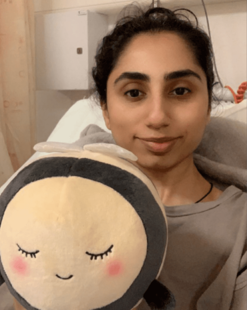 Mia is taking a selfie, sitting in a hospital bed. The selfie is cropped to show from the top of her head down to just below her chest. She is smiling slightly at the camera and holds a cuddly bee towards the camera.