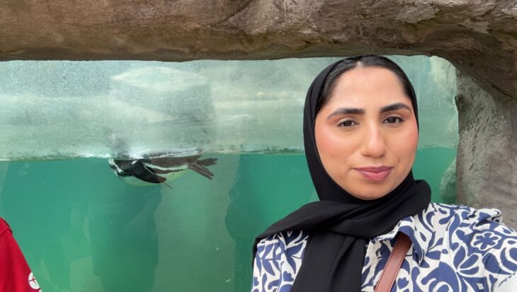 Mia is taking a selfie in front of an aquarium tank outside which contains a swimming penguin. She is smiling at the camera and wears a black hijab and blue and white patterned top. The photo is cropped to show her shoulders upwards.
