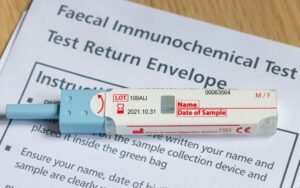 An unlabelled faecal immunochemical test sample tube lies on top of a sheet of paper that reads faecal immunochemical test test return envelope with instructions on top of a wooden table.