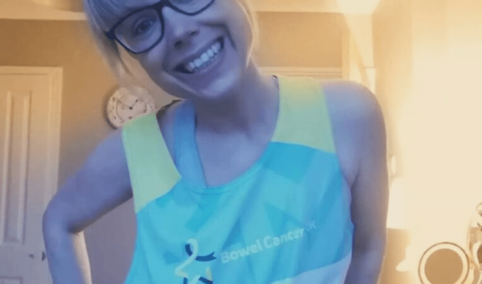 Rachel is grinning at the camera in a room in her house. She has her right hand on her hip and she is wearing the blue Bowel Cancer UK vest.
