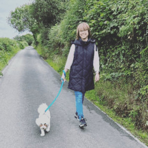 Rachel is smiling at the camera while walking her white dog down a country road with green bushes along the side.