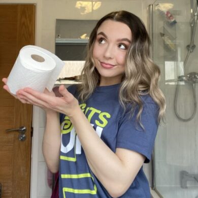 Abbie is standing in a white tiled bathroom. She is smiling facing the camera with her eyes staring to her right. She is wearing the navy Guts UK T-shirt and holding a toilet roll with both her hands.