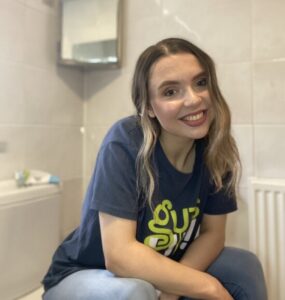 Abbie is in a bathroom and sat on top of the toilet with the seat closed. She is smiling at the camera with her hair down and wearing the navy Guts UK T-shirt with a pair of blue jeans. 