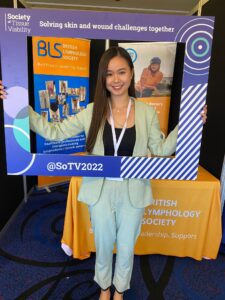 Dove is at a conference wearing a pastel green suit and holding a large frame in front of her that is from the society of tissue viability. Her upper half of her body is inside the frame. Behind her is an large orange banner and table labelled as the British Lymphology Society.