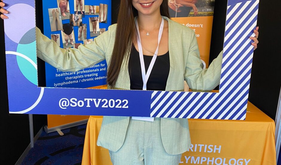 Dove is at a conference wearing a pastel green suit and holding a large frame in front of her that is from the society of tissue viability. Her upper half of her body is inside the frame. Behind her is an large orange banner and table labelled as the British Lymphology Society.
