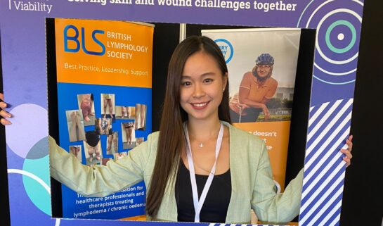 Dove is at a conference wearing a pastel green suit and holding a large frame in front of her that is from the society of tissue viability. Her upper half of her body is inside the frame. Behind her is an large orange banner and table labelled as the British Lymphology Society.