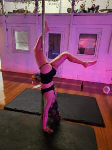 Dove is in a studio with a pole and crash mat in the centre of the room. She is doing a handstand with both hands on the crash mat and right leg hooked onto the pole and left leg behind her.