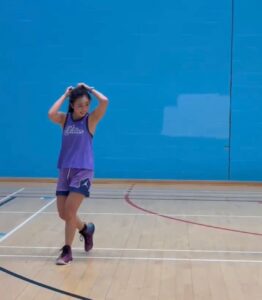 Dove is tightening her hairband at a basketball court. She is wearing purple basketball top and shorts and walking while staring towards her left.