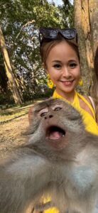 Dove is taking a selfie outdoors with a monkey. She is smiling at the camera with sunglasses on her head, wearing yellow earrings and dress. The monkey has their mouth open while looking into the camera.