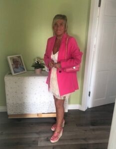 Lorna is stood indoors in front a green wall with a opened white door to her left. She is smiling at the camera in her wearing a white knee length dress, a hot pink long sleeved blazer, a crossbody bag and pink wedged sandals.
