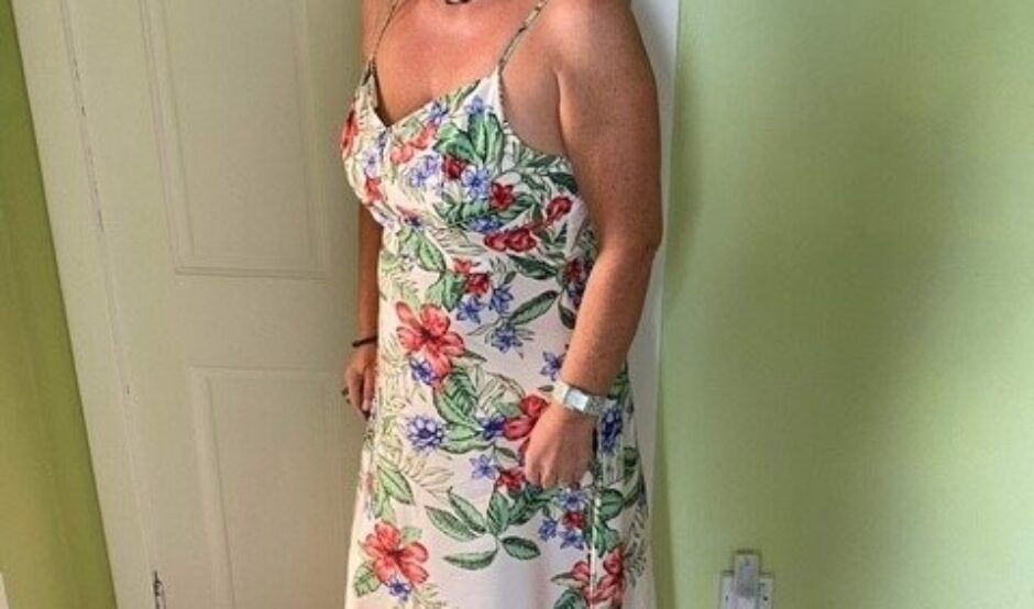 Lorna is stood in front a white wooden door and green walls. She is smiling at the camera with her blonde hair resting on her shoulder. She is wearing a long white summery floral dress and a navy flower fascinator hat.