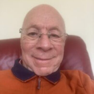 Peter is taking a selfie with him smiling in his orange long sleeved polo top. He is sat on his plush red couch.