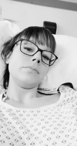A black and white selfie of Rachel who has taken it while she is laid on the hospital bed. She is wearing the hospital gown with her head on the pillow looking displeased. 