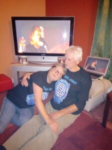 Diane is looking at the camera and resting peacefully beside her friend on the couch in her living room with orange and dark red walls. One of her arms is behind his back and the other lies on his lap. The TV is switched on behind them.