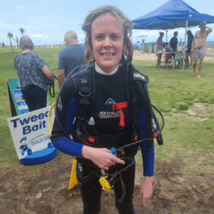 Sarah is seen wearing a scuba diving wet suit on a green field and smiling at the camera. She is stood on the grass with blue skies above her. There is a sign that says Tweed Bait behind her with a few other people stood behind her.