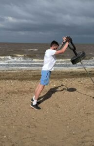 Ian is on the beach wearing a white top and blue shorts. He is leaning forwards and pushing some weights.