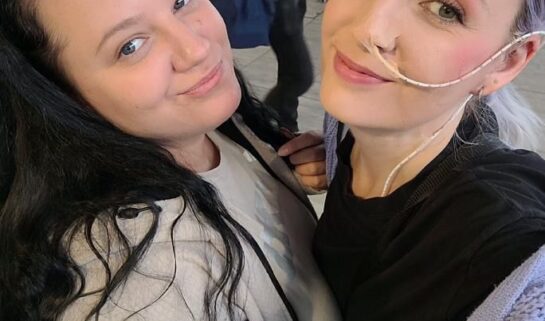 Leanne is taking a selfie with her friend and they are both smiling at the camera. Leanne is wearing a black top with a purple cardigan and he friend is wearing a beige jumper. Leanne has a tube passing through into nose.