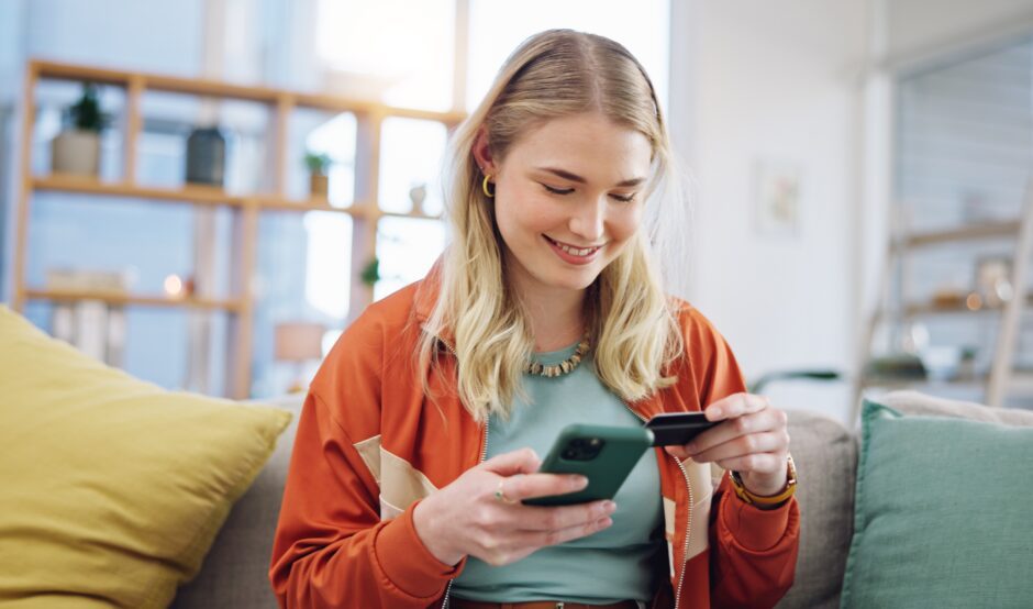 A woman browses online on her mobile phone whilst holding a bank card.