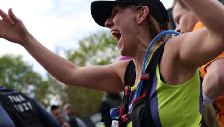 A person wearing a sports cap and a running vest has their hands up in the air, with a joyful expression on their face as they run amongst a crowd of runners.