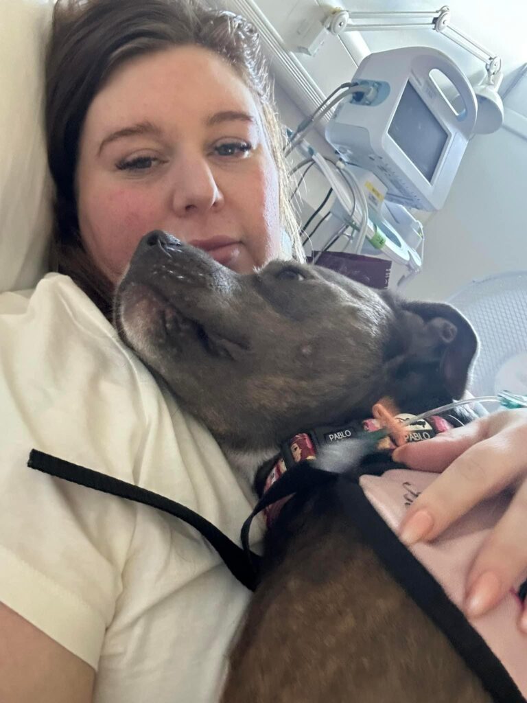 Amee is lying in a hospital bed with various monitoring machines around her. Her assistance dog, Belle, is resting on her chest.