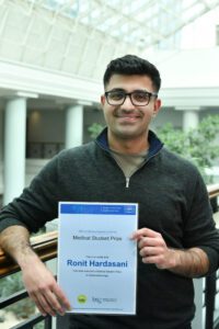 Ronit holds his awards certificate and is smiling at the camera