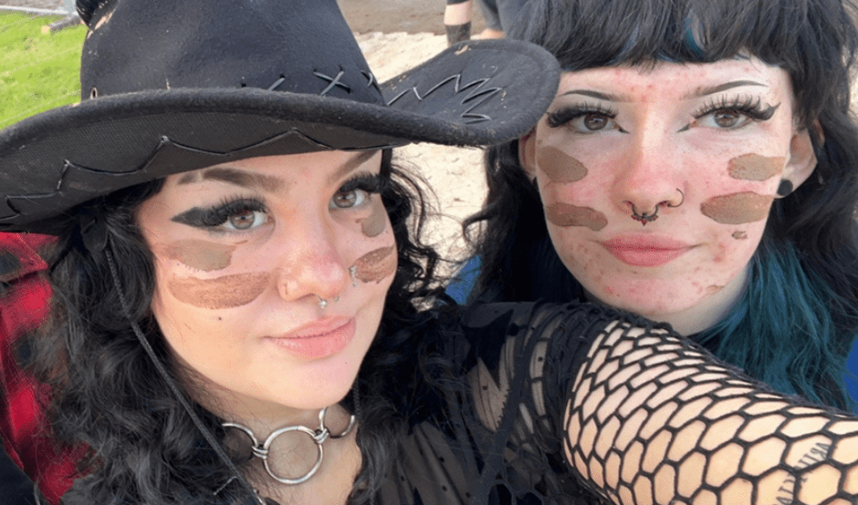 Rachel (left) wears a black cowboy hat, with fishnet style sleeves and a top and has black, long, curly hair. She is smiling at the camera and has two stripes of brown face paint on each of her cheeks. She is with her friend at a music festival.