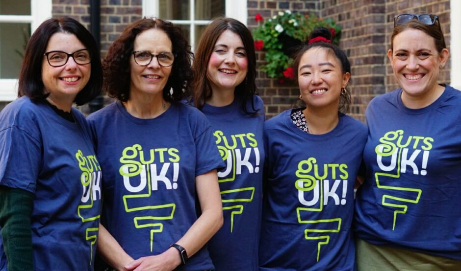 Cherylyn, Melanie, Amy, Leeona and Katie are smiling at the camera wearing Guts UK t-shirts. There is brick wall with white framed windows behind them.