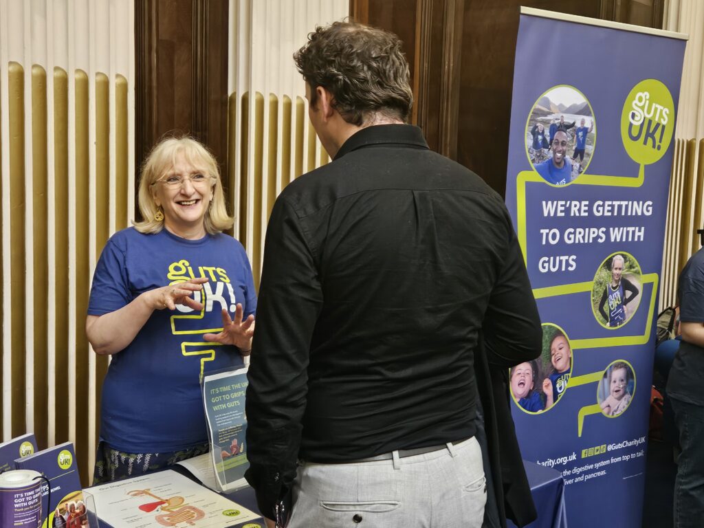 A member of the Guts UK is standing behind a table with information leaflets. They are talking to a person on the other side of the table, who has their back to the camera.