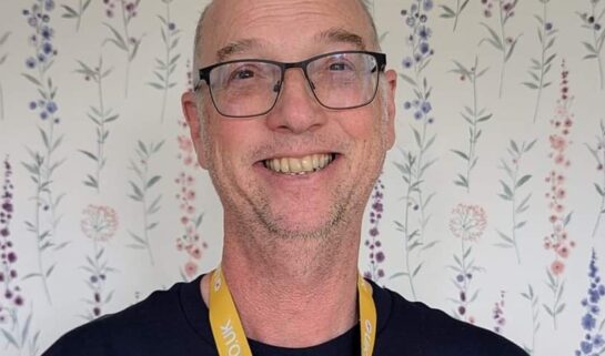 Stephen stands in front of a white wallpaper background, with delicate, floral detailing on it in different colours. It is a headshot of Stephen, and he wears black, rectangular glasses with curved edges, a black t-shirt and a yellow lanyard. He is smiling at the camera.